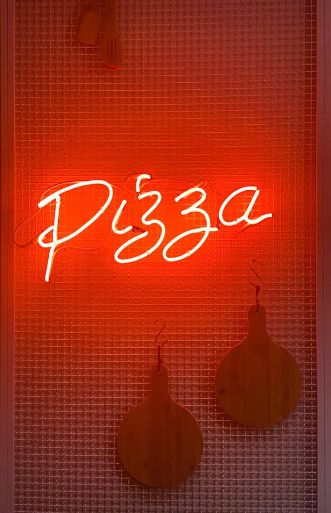 A glowing neon sign displaying the word Pizza on a textured red background.